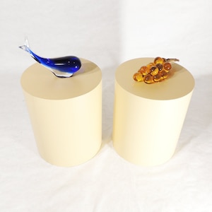 May include: Two cream-colored cylindrical pedestals, one with a blue glass whale sculpture and the other with a cluster of amber glass grapes.