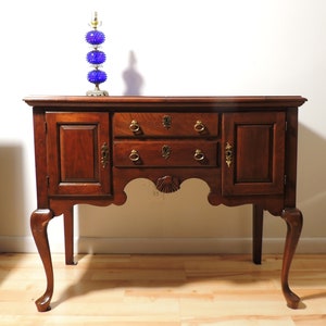 May include: A mahogany wood sideboard with two doors and three drawers. The sideboard has cabriole legs and a carved shell design on the apron. A blue glass lamp sits on top of the sideboard.