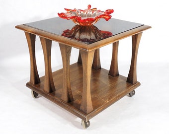 May include: A mid-century modern end table with a dark wood finish and a glass top. The table has four legs with a unique curved design and four wheels. A red glass bowl sits on the table top.