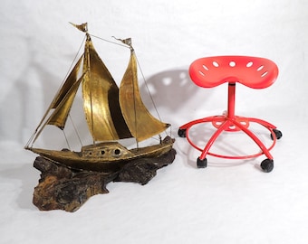 Pode incluir: Uma escultura de veleiro de metal dourado com uma base de madeira. O veleiro tem três velas e está sentado sobre uma superfície branca. Uma cadeira giratória vermelha com um assento redondo está no fundo.