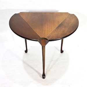 Op de afbeelding: Een donkerbruine, halfronde tafel met een uniek ontwerp. Het tafelblad is verdeeld in drie secties met verschillende houtnerfpatronen. Hij heeft drie slanke, donkere poten, elk ongeveer 71 cm lang, met een afgeronde voet.