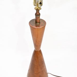 Puede incluir: Una lámpara de mesa moderna de mediados de siglo con una base de madera en forma de reloj de arena. La lámpara tiene un casquillo de latón y cobre y un cable eléctrico marrón. La lámpara está sobre un fondo blanco.