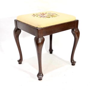 Peut inclure: Un petit tabouret en bois avec un coussin à motif floral. Le tabouret a quatre pieds cabriole et un dessus rectangulaire.