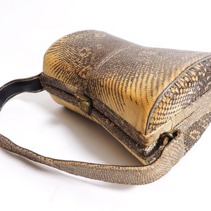 Könnte beinhalten: Vintage Handtasche mit Reptilienhautmuster in Beige- und Brauntönen. Die Tasche hat eine abgerundete Form mit einem strukturierten Rahmen, einem oberen Griff und einem Metallverschluss. Der Riemen ist ca. 76 cm lang.