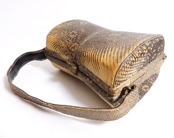 Könnte beinhalten: Vintage Handtasche mit Reptilienhautmuster in Beige- und Brauntönen. Die Tasche hat eine abgerundete Form mit einem strukturierten Rahmen, einem oberen Griff und einem Metallverschluss. Der Riemen ist ca. 76 cm lang.
