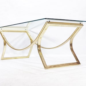 May include: A glass-top coffee table with a brass frame. The frame is made of two curved pieces of brass that meet in the middle. The table has a modern design.