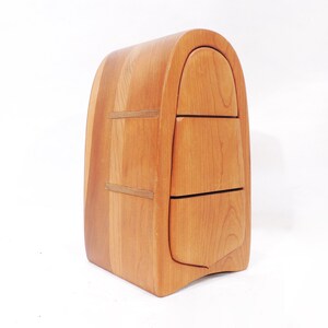 Pu&ograve; includere: Uno scrigno in legno con tre cassetti, ognuno con un fronte curvo. Lo scrigno &egrave; fatto di legno marrone chiaro e ha una parte superiore arrotondata.