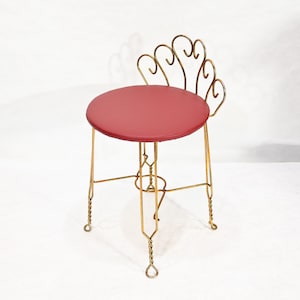 Può includere: Sgabello da toeletta vintage con seduta rotonda rossa e struttura decorativa in metallo color oro. Lo schienale dello sgabello presenta un design a forma di ventaglio. Le gambe hanno un design a corda intrecciata.