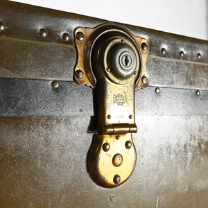 May include: Close-up of a vintage metal suitcase with a brass latch and lock. The suitcase has a black leather trim and is secured with rivets. The lock has the inscription "Excelsior" and "Made in USA".