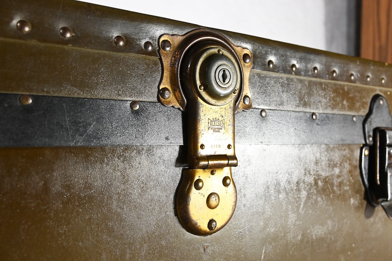 May include: Close-up of a vintage metal suitcase with a brass latch and lock. The suitcase has a black leather trim and is secured with rivets. The lock has the inscription "Excelsior" and "Made in USA".