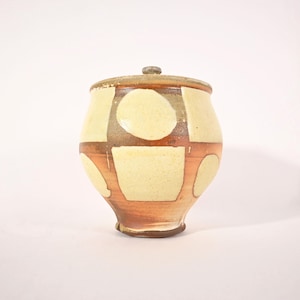 Könnte beinhalten: Keramiktopf mit Deckel, mit einem cremefarbenen und braunen geometrischen Muster. Der Topf hat einen abgerundeten Körper und einen kleinen Knopf auf dem Deckel. Das Design umfasst Quadrate und Kreise mit einer rötlich-braunen Akzentfarbe.