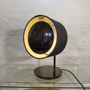 May include: A black and white table lamp with a round, fabric shade. The shade has a yellow inner lining and a black outer lining. The lamp is on a glass table.