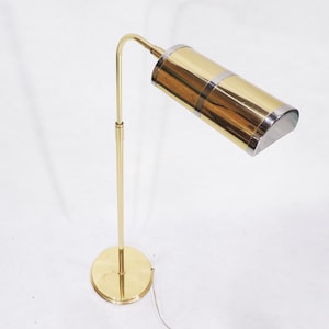 May include: A gold floor lamp with a curved arm and a cylindrical shade. The shade is made of brass and has a silver trim.