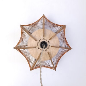 以下が含まれることがあります： 星型のデザインの茶色とベージュのペンダントライト。ライトは「SAVELI ST. PAUL MN」の文字が入った木製のベースと、紐のような織り方のシェードが付いています。ベージュ色のコードが中央から吊り下がっています。