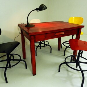 May include: A red wooden table with a drawer, a black lamp with a white shade, and three chairs. Two of the chairs are black and one is yellow. The table is surrounded by a white wall.