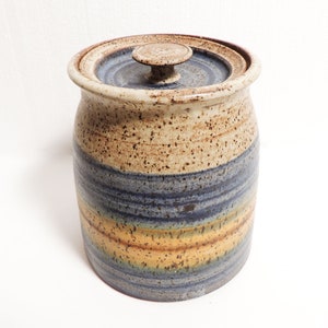 Può includere: Un vaso di ceramica marrone e blu con un coperchio. Il vaso ha una glassa macchiata e una fascia di strisce blu e gialle intorno al centro.