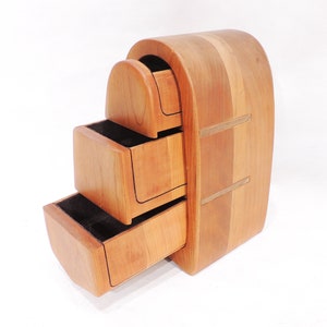 Pu&ograve; includere: Una scatola di legno con tre cassetti, ognuno con un fronte curvo. La scatola &egrave; realizzata in legno chiaro e scuro, e i cassetti sono rivestiti in feltro nero. La scatola &egrave; leggermente aperta, con il cassetto inferiore tirato fuori.