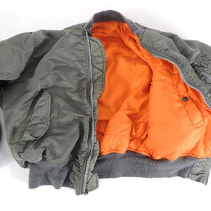 May include: An olive green bomber jacket with an orange lining. The jacket features a zip closure, two front pockets, and ribbed cuffs and hem. A classic military-style outerwear garment.