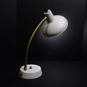 May include: A gray desk lamp with a flexible arm and a white shade. The lamp has a gold-colored base and a black knob on the top.