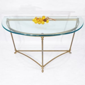Puede incluir: Una mesa consola de media luna con una tapa de vidrio y un marco de latón. La mesa tiene una tapa de vidrio curvada y una base triangular. La mesa está decorada con un grupo de bolas de vidrio ámbar.