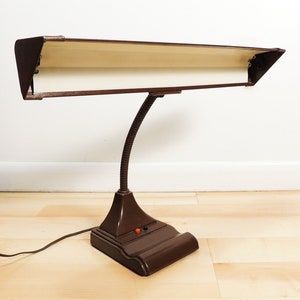 May include: A brown vintage desk lamp with a flexible arm and a long, rectangular shade. The lamp has a white interior and a black cord.