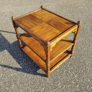 May include: A three-tiered wooden side table with a brown finish. The table has a square top and is supported by four turned legs. The top shelf has a parquet design.