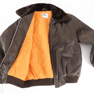 May include: A brown bomber jacket with a faux fur collar and an orange quilted lining. The jacket has a zipper closure, a ribbed hem and cuffs, and a small pocket on the sleeve. A tag is visible on the bottom left.