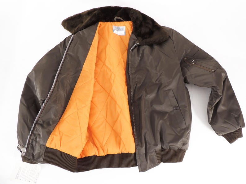 May include: A brown bomber jacket with a faux fur collar and an orange quilted lining. The jacket has a zipper closure, a ribbed hem and cuffs, and a small pocket on the sleeve. A tag is visible on the bottom left.