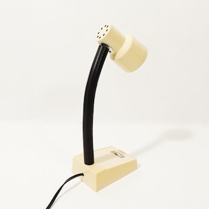 Op de afbeelding: Een beige bureaulamp met een flexibele zwarte nek en een ronde kap. De lamp heeft een eenvoudig ontwerp en is aangesloten op een stopcontact.