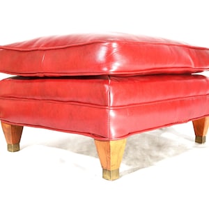 May include: A square ottoman upholstered in bright red leather with a thick, tufted top cushion. The base features four tapered light-colored wooden legs, each finished with a square metallic cap at the bottom, providing a stable foundation.