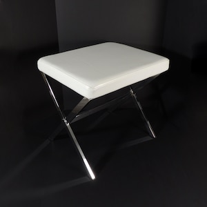 May include: A white, square ottoman with a padded top and a polished chrome X-shaped base. The ottoman is set against a black background, highlighting its modern design. The clean lines and neutral color palette make it a versatile piece of furniture.