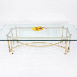 May include: A rectangular glass coffee table with a brass frame. The frame has a unique design with curved supports and decorative accents. The table is sitting on a white surface.