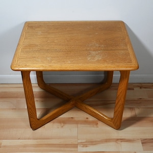 May include: A square wooden coffee table with a light brown finish. The table has a simple, modern design with a cross-shaped base.