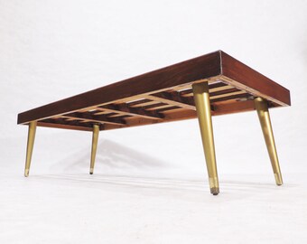 May include: A long, rectangular wooden bench with a slatted top and four gold-colored legs. The bench is made of dark wood and has a simple, mid-century modern design.