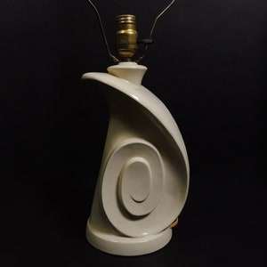 May include: A white ceramic table lamp with a swirling, abstract design. The lamp has a simple, round base and a gold-colored socket.