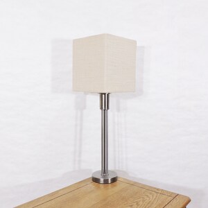 May include: A silver table lamp with a square, off-white linen lampshade. The lamp has a simple, modern design.