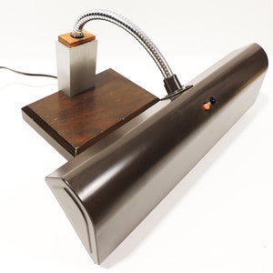 Op de afbeelding: Een bruine metalen bureaulamp met een flexibele arm en een houten voet. De lamp heeft een zilveren schakelaar en een bruin snoer.