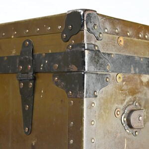 May include: Close-up of a vintage green metal trunk with black metal hardware, including hinges, corner protectors, and a latch. The trunk has a worn, distressed look, with visible rivets and scratches.