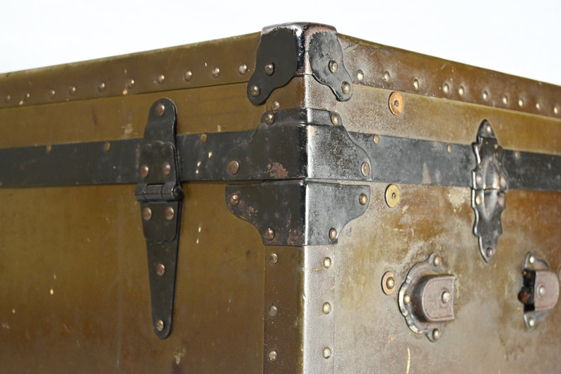 May include: Close-up of a vintage green metal trunk with black metal hardware, including hinges, corner protectors, and a latch. The trunk has a worn, distressed look, with visible rivets and scratches.