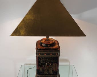 May include: A decorative table lamp with a square lampshade and a base made from a vintage tea tin. The lamp features a black and red painted design with figures and a gold-colored lampshade. The lamp sits on a wooden base.