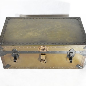 May include: A vintage metal storage trunk with a brass finish and black accents. The trunk has two latches and a lock on the front.