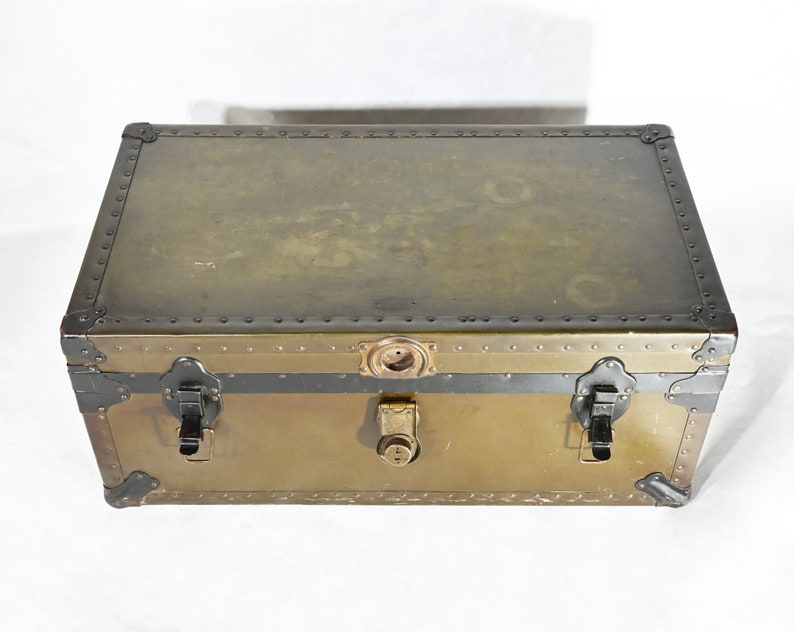 May include: A vintage metal storage trunk with a brass finish and black accents. The trunk has two latches and a lock on the front.