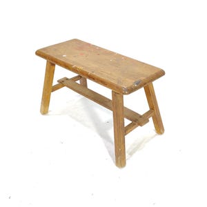 May include: A simple wooden stool with a rectangular seat and a single stretcher underneath. The stool is made of light brown wood and has a rustic, vintage look.