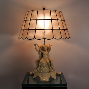 May include: A table lamp with a white and black lampshade and a base featuring three cherubs. The lampshade is made of a translucent material with a scalloped edge.