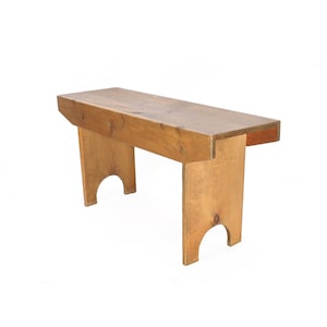 May include: A simple wooden bench with a natural finish. The bench has a rectangular top and four legs. The legs are slightly curved at the bottom.