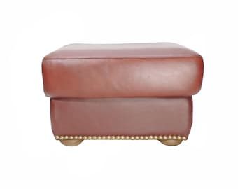 Sillón otomano de cuero burdeos, taburete de color intenso con cojín superior y detalles de tachuelas de latón.