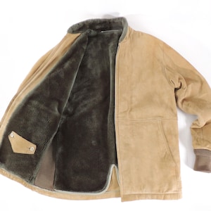 Op de afbeelding: Een bruin suède jasje met een donkerbruine bontvoering. De jas heeft een ritssluiting, een bruine geribde manchet en een klein zakje met een knoopsluiting. De kraag is gevoerd met donkerbruin bont.