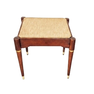 May include: A small, square wooden stool with a woven beige seat. The dark brown legs are tapered and have gold-colored accents. The stool is approximately 18 inches tall.