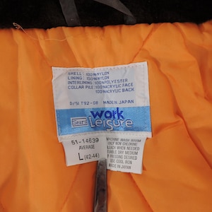 May include: Close-up of a vintage jacket's interior, featuring a bright orange quilted lining and a white tag with text. The tag indicates the jacket is a size L (42-44). The jacket's shell is 100% nylon.