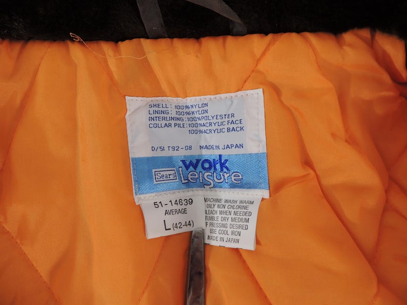 May include: Close-up of a vintage jacket's interior, featuring a bright orange quilted lining and a white tag with text. The tag indicates the jacket is a size L (42-44). The jacket's shell is 100% nylon.